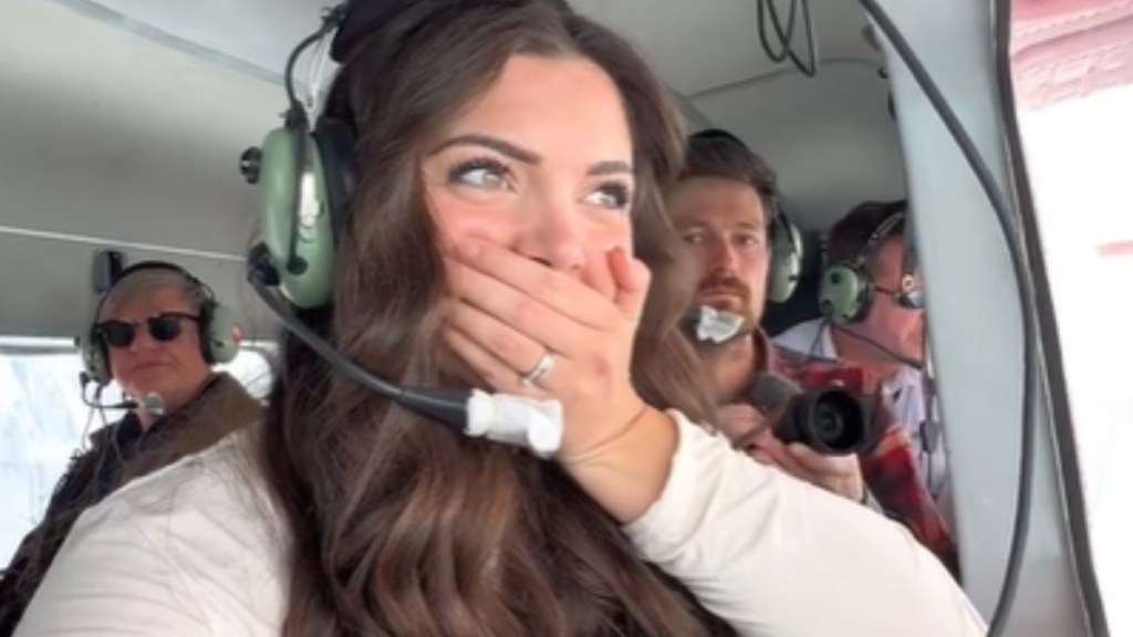 Close up of a woman on a plane. She's wearing a headset and is looking out of the window. Her eyes are wide in amazement and she covers her mouth with her hand.