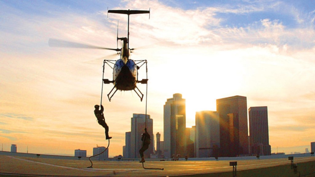 A helicopter flies close to the rooftop of a building. Two people hold onto ropes attached to the helicopter. The sun is setting in the distance