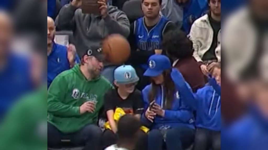 A dad, mom, and kid sit together in front-row seats at a basketball game. They're looking at each other as a basketball is headed straight toward the little kid who sits in the middle