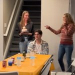 A man sits at a dining table, smiling as he waits. Mom and Dad stand to the side, happily waiting for their son's girlfriend to place the cake on the table. The girlfriend holds the cake, starting to walk around to place it on the table.