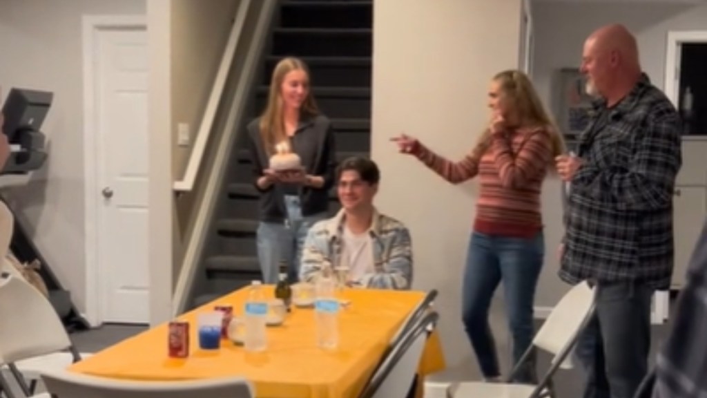 A man sits at a dining table, smiling as he waits. Mom and Dad stand to the side, happily waiting for their son's girlfriend to place the cake on the table. The girlfriend holds the cake, starting to walk around to place it on the table.