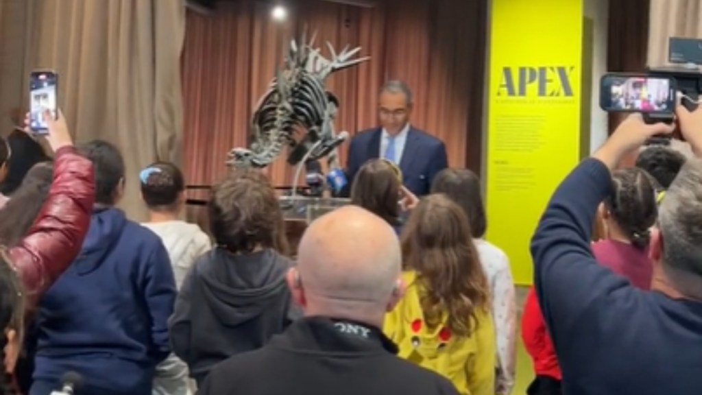 A crowd watches on as a man speaks at a podium in front of a newly unveiled dinosaur specimen at the American Museum of Natural History