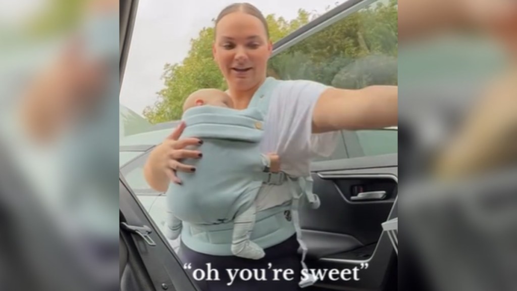 A woman stands at her car, door open, as she puts food inside. Her baby is strapped to her chest. She smiles. Text on the image shows the woman saying: "oh, you're sweet"