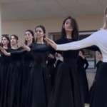 A line of women hold their arms out elegantly as they perform a traditional North Caucasus dance. They're wearing all black shirts and long, flowy skirts except the woman in the lead who wears a white shirt