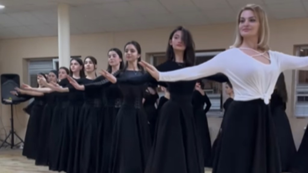 A line of women hold their arms out elegantly as they perform a traditional North Caucasus dance. They're wearing all black shirts and long, flowy skirts except the woman in the lead who wears a white shirt