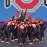 A group of dancers gather in a circle, lifting a single dancer above them. This single dancer holds out her arms and holds her head back, seemingly screaming