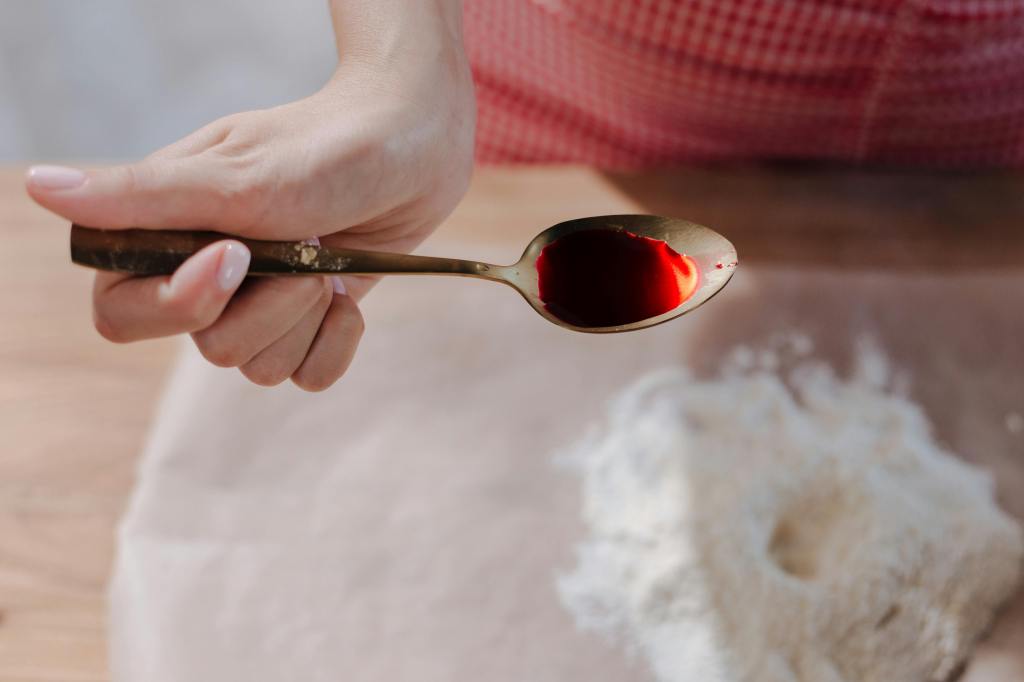 Top-down view of a spoon that's holding red dye. The spoon is being held by a woman. Below on the table is flour. 