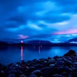 Sunset in Alaska. The sky and image overall has an incredibly vibrant blue hue. The sun is setting over a body water. A shore of rocks are near the camera and, at a distance, there are mountains