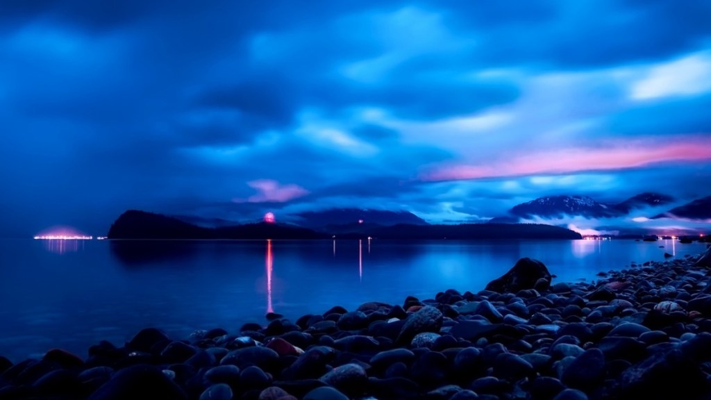 Sunset in Alaska. The sky and image overall has an incredibly vibrant blue hue. The sun is setting over a body water. A shore of rocks are near the camera and, at a distance, there are mountains