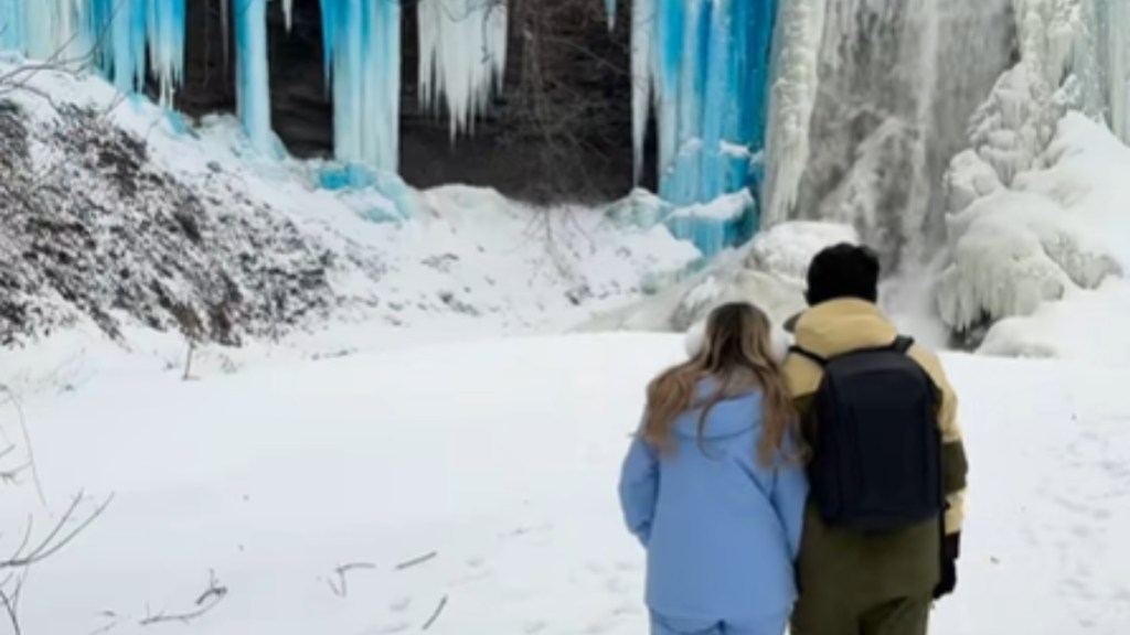 View from behind of a man and woman walking in the snow toward a waterfall in the distance. The water at the falls is frozen. Many of the icicles are a vivid blue color.