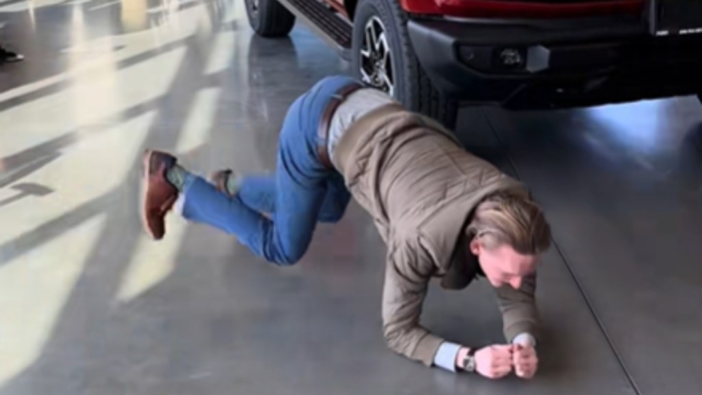 A man crawls on his arms, one foot raised slightly in the air. Just behind him, a red vehicle can be partially seen