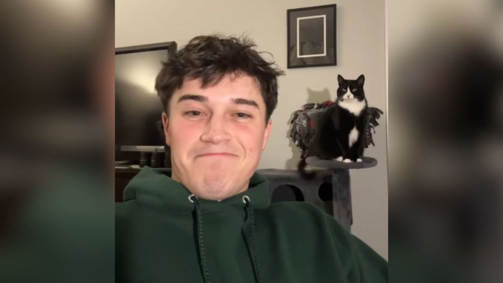 A man smiling while his cat sits in the background.