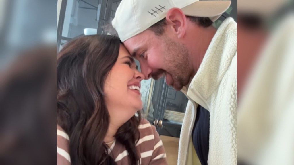 Close up of a man and woman. They stand close, face to face, with their foreheads and noses touching. The woman laughs and smiles as the man smiles and talks