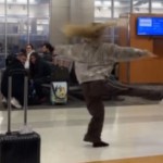 The interior of an airport. Most people are sitting, but one woman is in the aisle, dancing. She has one leg and both arms raised as she spins, hair flying in the air