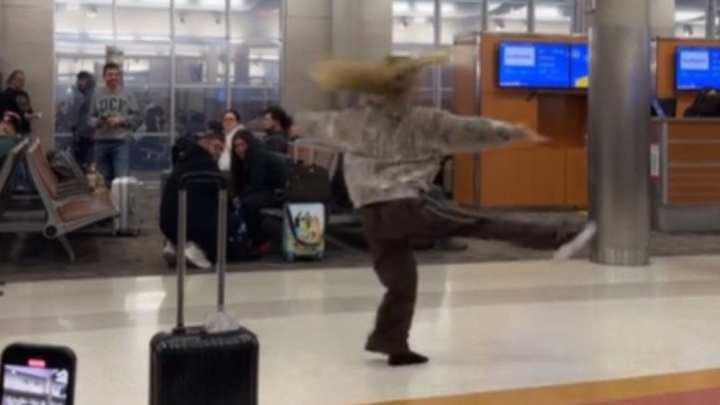 The interior of an airport. Most people are sitting, but one woman is in the aisle, dancing. She has one leg and both arms raised as she spins, hair flying in the air