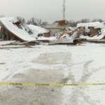 View of a collapsed fire station from behind yellow tape placed to keep people out. Snow is covering the ground and the collapsed building