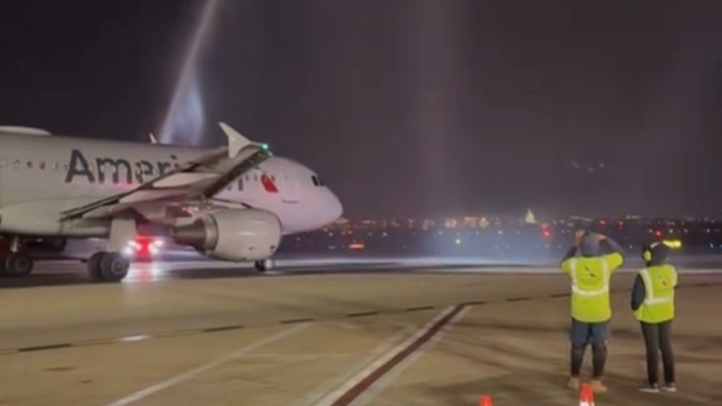 At a distance, an American Airlines plane travels across the ground after landing. It's approaching water being shot into the sky. Two airline workers stand nearby and watch. It's night, so the city lights shine at a distance