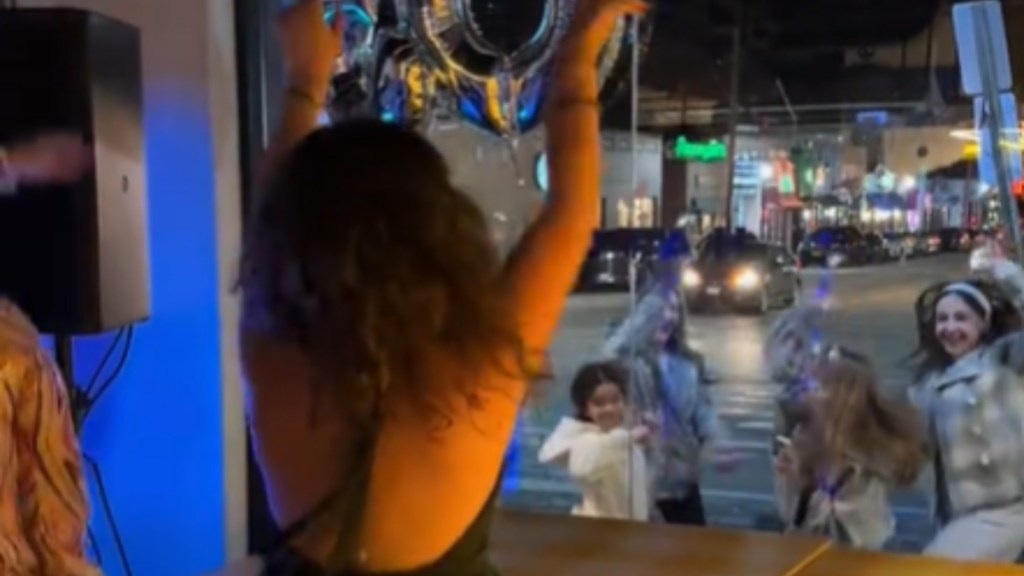 View from behind of a woman dancing, arms raised in the air. Outside, a group of young girls can be seen dancing with her