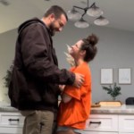 A man and woman stand in a kitchen, facing one another. The man is tall and the woman is short, so he looks down as he rests his hands on her arms. He smiles widely at her and she smiles back