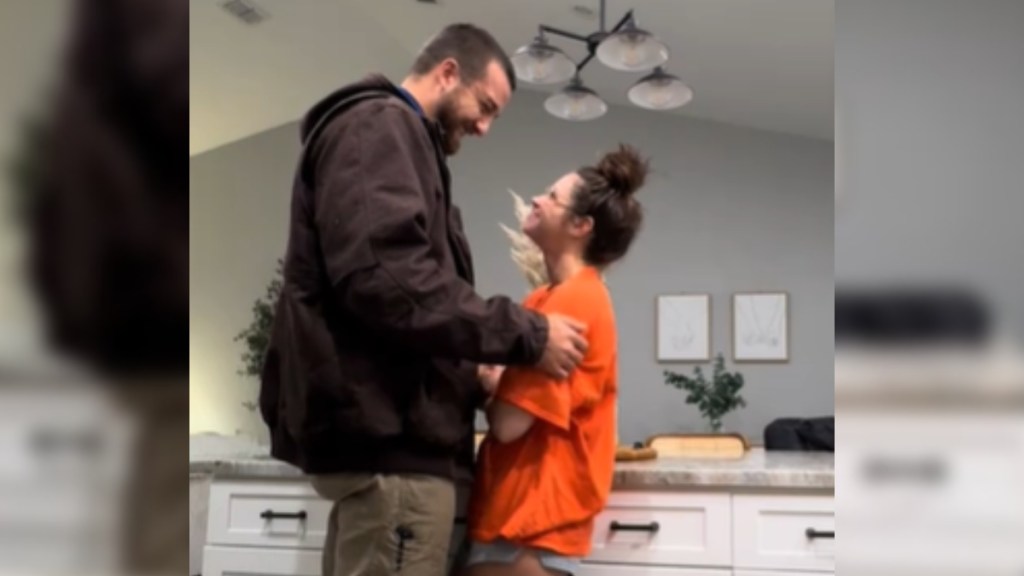 A man and woman stand in a kitchen, facing one another. The man is tall and the woman is short, so he looks down as he rests his hands on her arms. He smiles widely at her and she smiles back