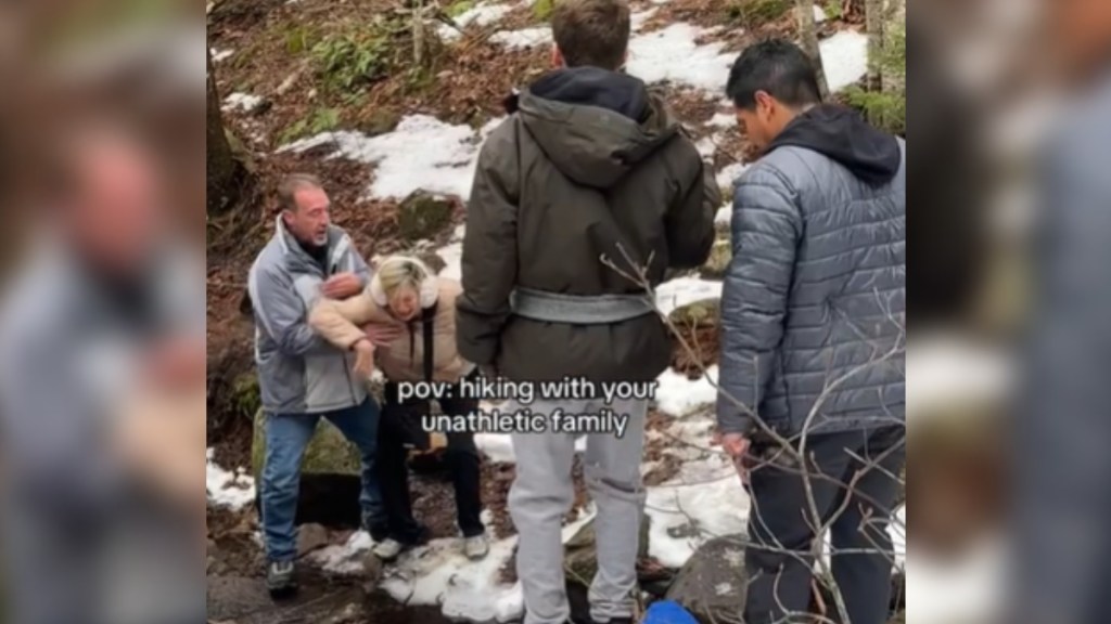 Two men stand near each other, looking on at Mom and Dad. Dad helps Mom who is struggling to walk over a small creek. She looks distressed. Text on the image reads: POV: Hiking With Your Unathletic Family