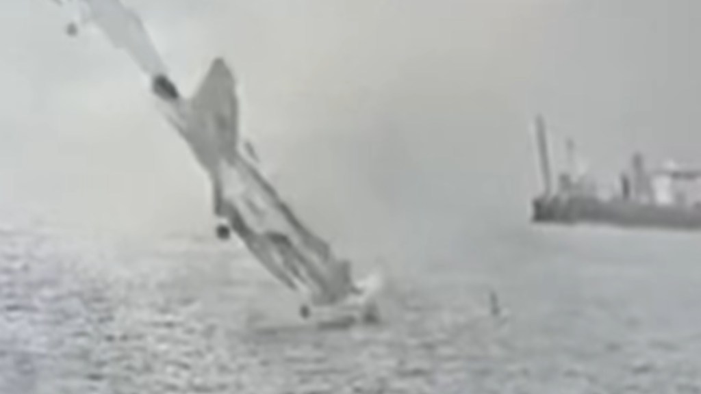 A fighter jet starts to crash, nose first, into water. A nearby fishing vessel can be seen at a distance