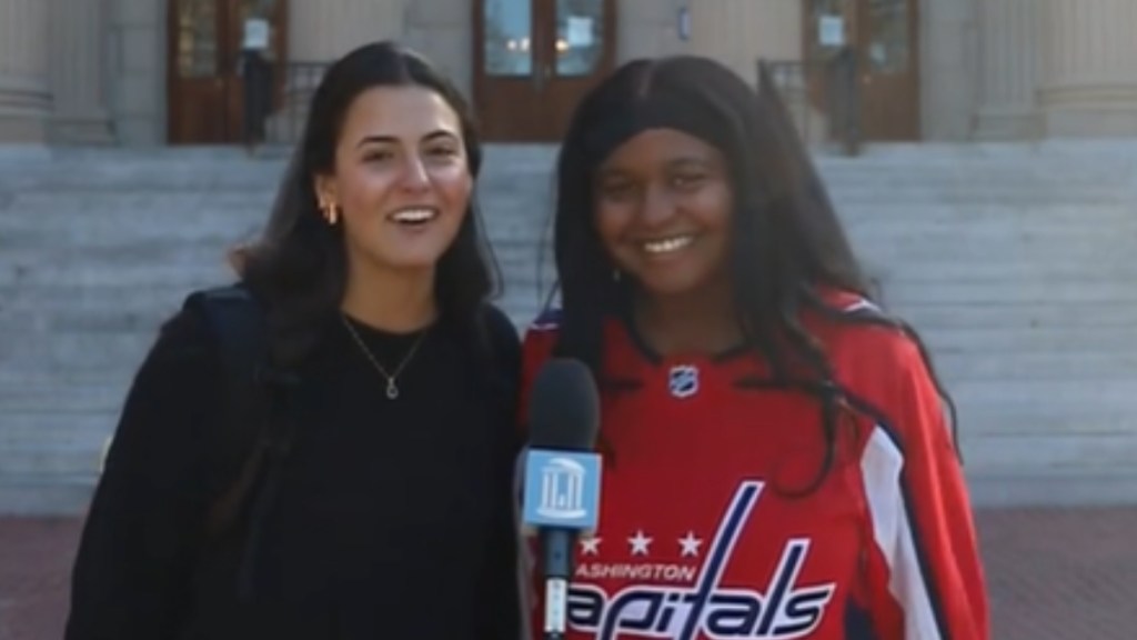 Two young women stand outside and in front of a mic placed on a stand. One woman smiles, teeth showing. The other smiles as she talks