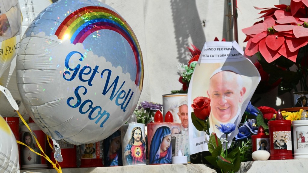 Close up of photos, candles, and a balloon left outside for Pope Francis as he recovers from an illness in the hospital. The balloon says "get well soon" and has a rainbow on it