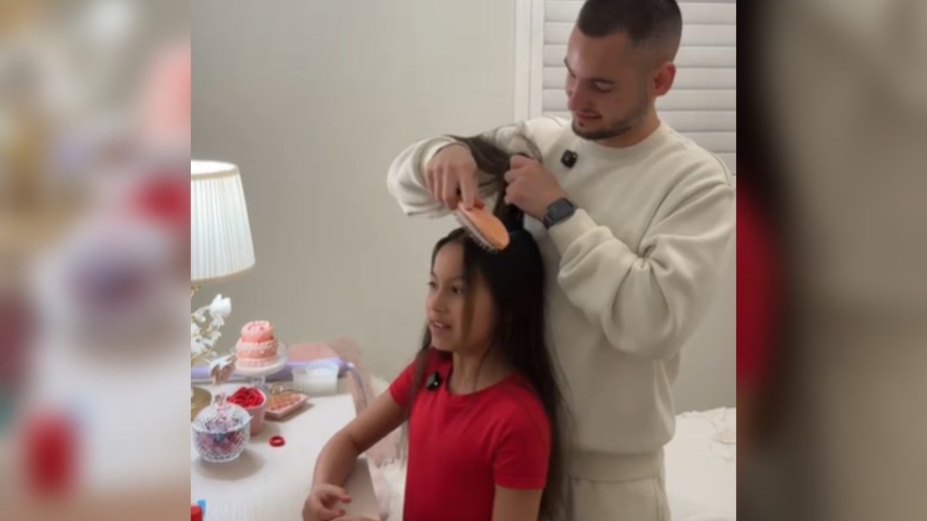 A dad and daughter stand in front of a dresser with a mirror. Dad smiles as he pulls back a section of the girl's hair, styling it. She stands still, smiling as she looks in the mirror that can't be seen in this image