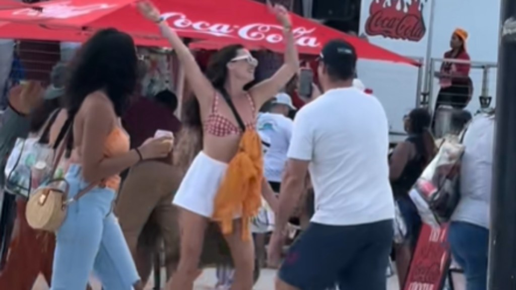 From a distance, a man records a woman dancing with her hands raised in the air. Another woman walks past, looking at them