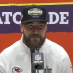 Travis Kelce sits in front of a mic for media interviews. He smiles