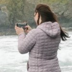 Close up of a woman standing on the shore. Her back is mainly toward the camera. She has her phone out and is taking a photo of something closer to the water