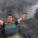 A man rests on the side of a summit, hands in the air in celebration. In the distance, the debris and smoke of volcano eruption can be seen