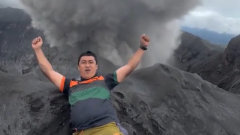 A man rests on the side of a summit, hands in the air in celebration. In the distance, the debris and smoke of volcano eruption can be seen