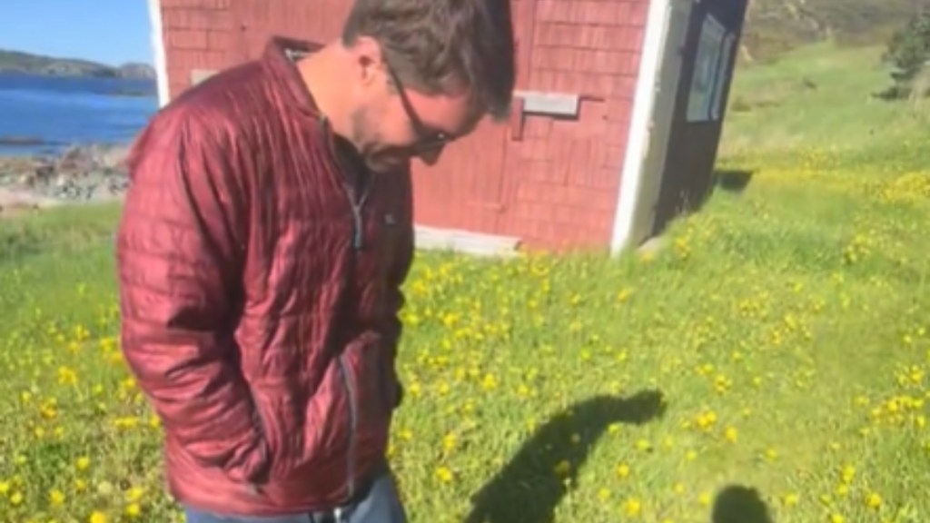 A man stands outside in bright green grass with tons of yellow flowers. A red building is just behind him. He smiles, looking down at something that can't be seen in this image. His shadow, and the shadow of someone else, can be seen on the ground