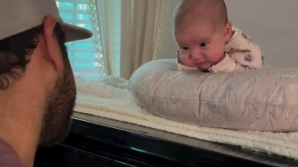 Close up of a man sitting at a piano, playing. On top of the piano is a baby resting on her tummy on something comfy. She rests her chin on her tiny fists, looking closely at dad.