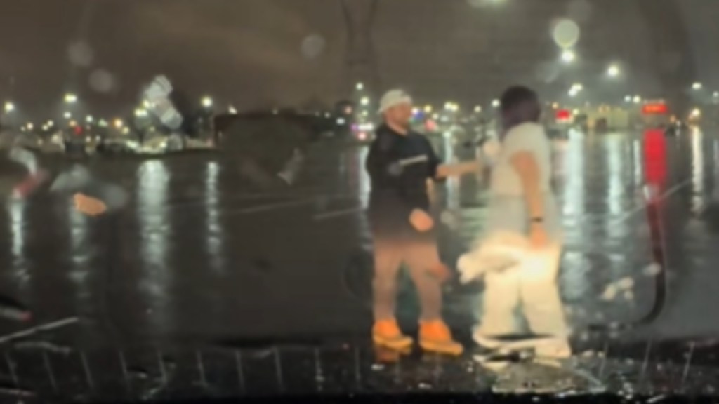 View of a parking lot as seen from a parked vehicle. A man takes a woman's hand, leading her toward him. It's night and raining. Lights from other cars and buildings can be seen in the distance