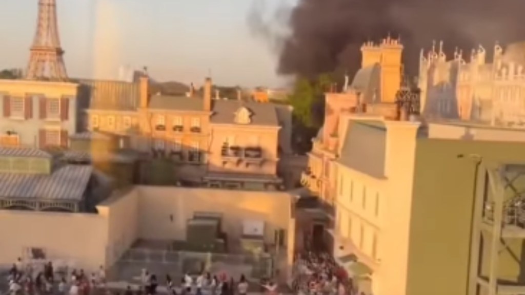 Aerial view of Disney's EPCOT. People gather on the ground while a giant plume of black smoke rises in the distance