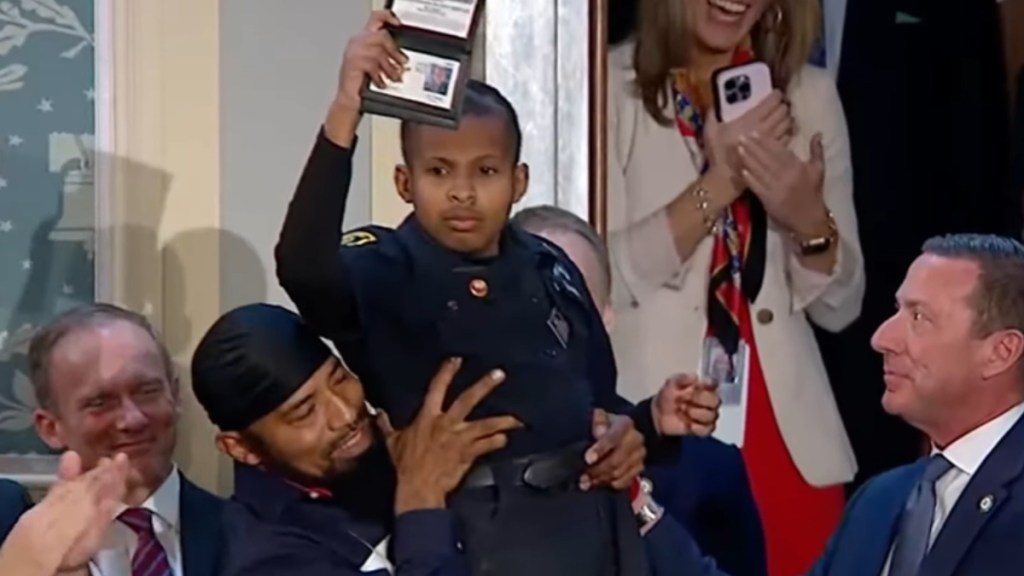 A man smiles as he holds up his 13-year-old son, DJ Daniel, into the air. The teen is wearing a police uniform and is holding an ID badge into the air. A number of government workers and politicians stand or sit nearby and cheer