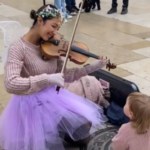 A woman and toddler rest on the ground outside. The woman wears a purple tule skirt, flower crown, and fairy wings while playing a violin. She's smiling. The toddler wears a similar sweater to the woman and looks up at her, entranced