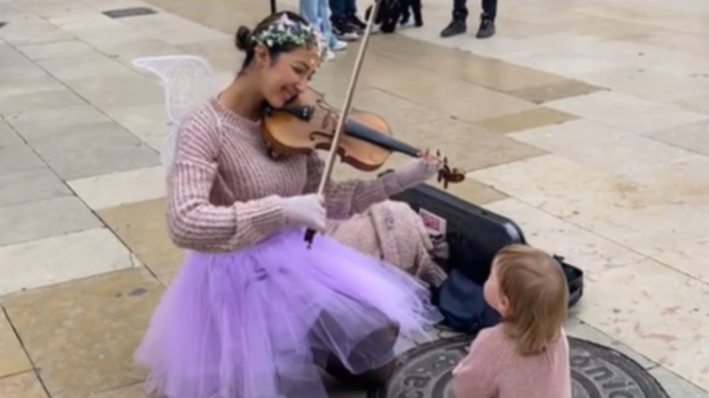 A woman and toddler rest on the ground outside. The woman wears a purple tule skirt, flower crown, and fairy wings while playing a violin. She's smiling. The toddler wears a similar sweater to the woman and looks up at her, entranced