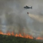 Distant view of an intense amount of smoke rising from a line of fires in a grassy area. A helicopter flies nearby, dropping water on the flames