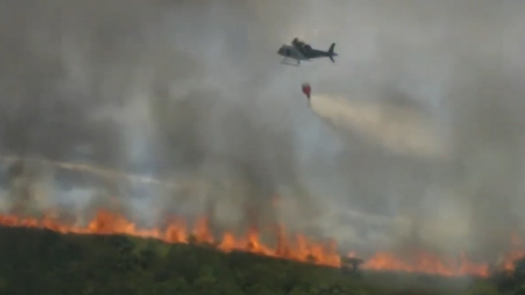 Distant view of an intense amount of smoke rising from a line of fires in a grassy area. A helicopter flies nearby, dropping water on the flames