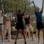 A group of five people stand in a formation, dancing. Everyone has their hands raised in the air. They're dancing outside, trees at a distance behind them