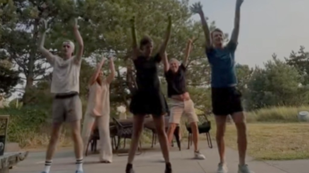 A group of five people stand in a formation, dancing. Everyone has their hands raised in the air. They're dancing outside, trees at a distance behind them