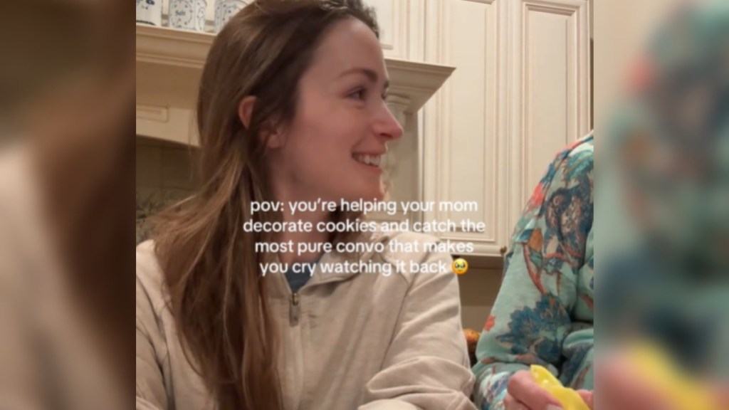 Close up of a woman smiling as she looks off to the side. In this image, the arm of a woman standing next to her can be seen. Text on the image reads: "Pov: you’re helping your mom decorate cookies and catch the most pure convo that makes you cry watching it back"