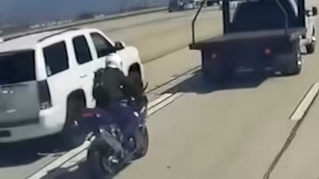 Dashcam footage of a Los Angeles freeway. In one lane, there's a white vehicle. Driving around the same speed in the lane over is a motorcyclist. Not too far in front of the motorcycle is a truck with a large bed.