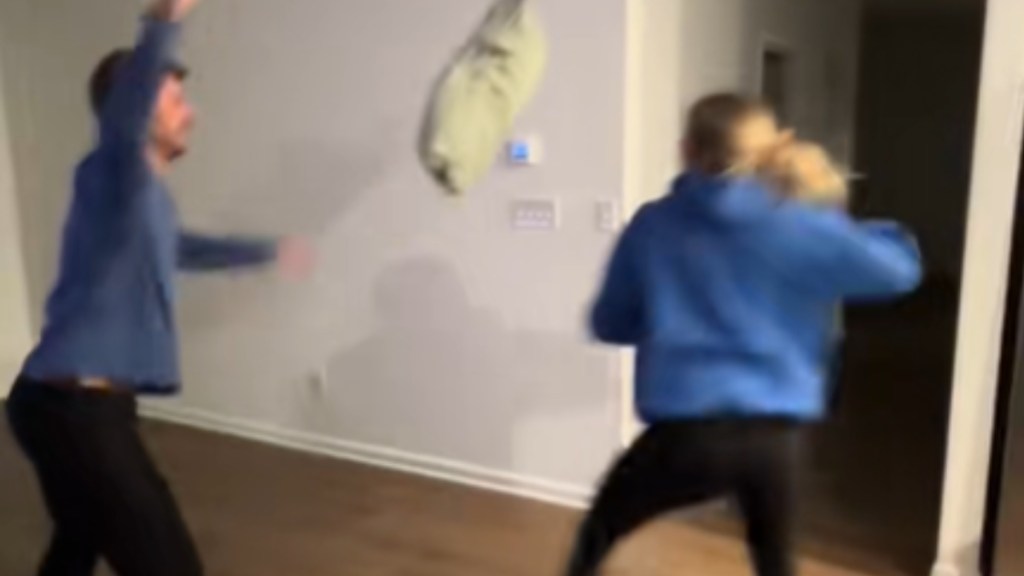 A man has his arms raised from tossing the pillow that's currently mid-air. A woman stands across from the pillow, stance indicating she's about to punch it