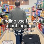 View from behind of a man walking down the aisle of a grocery store. He's dragging two rolling suitcases, one in each hand. Text on the image reads: Finding use for your giant luggage (now that they don't fly for free)