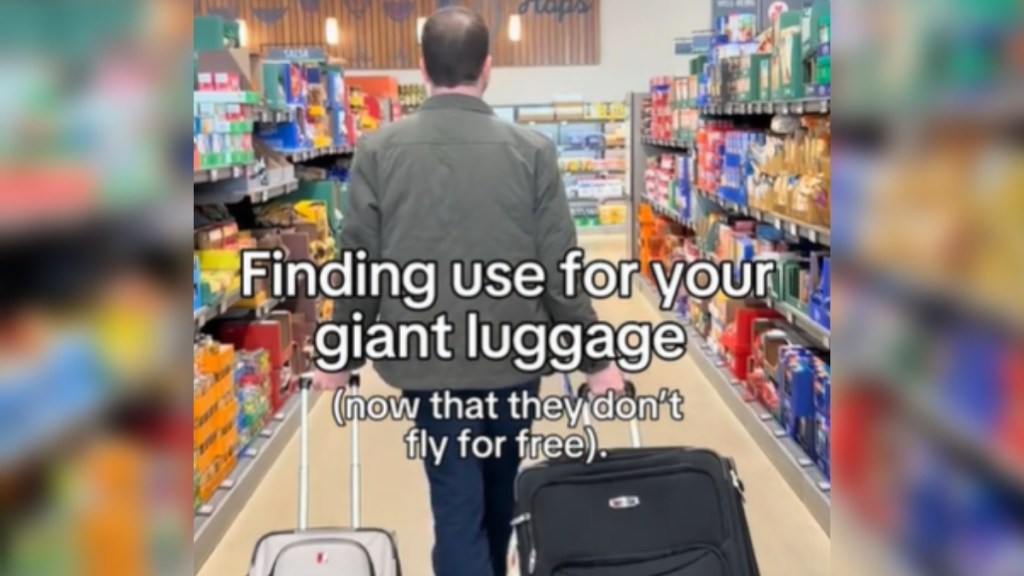 View from behind of a man walking down the aisle of a grocery store. He's dragging two rolling suitcases, one in each hand. Text on the image reads: Finding use for your giant luggage (now that they don't fly for free)